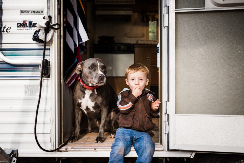A little boy sitting in the doorway of an RV eating something with his dog sitting next to him. 